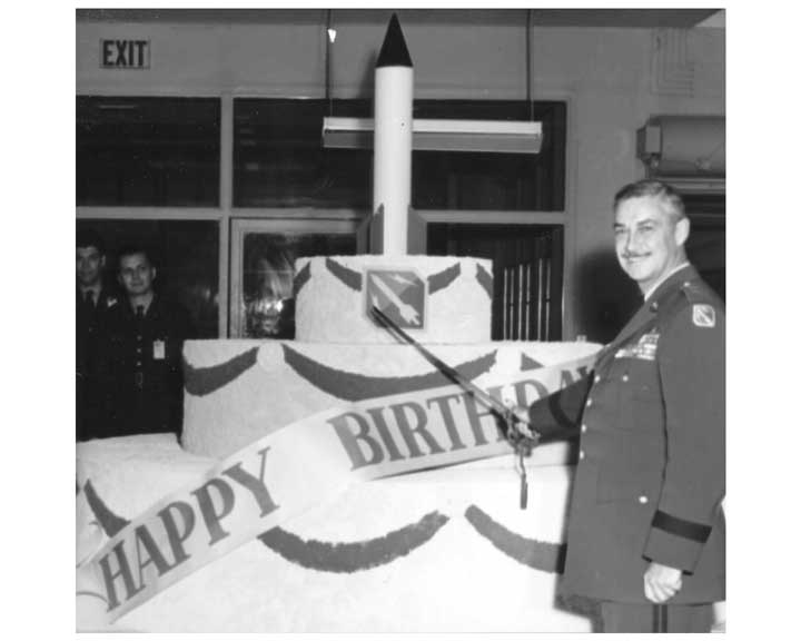 Photo of an officer in dress uniform, pretending to cut a large rocket cake with a saber for the Army Ballistic Missile Agency's first birthday.  The cake has a banner that says HAPPY BIRTHDAY.