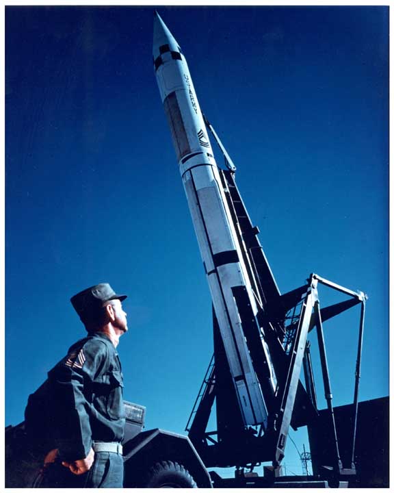 Photo of SERGEANT short-range, surface-to-surface missile being raised in to firing position on its trailer launcher, an in the foreground an Army master sergeant in fatigues watching the missile being raised.