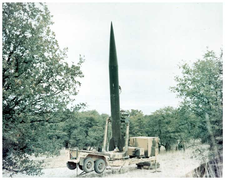 Photo of SERGEANT short-range, surface-to-surface missile in its raised firing position on its trailer launcher in a wooded area.